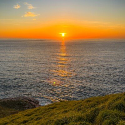 the sun is setting over the ocean on a grassy hill